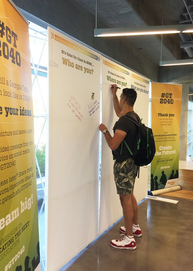 A tall male student wearing shorts and a backpack stands before three eight-foot-high panels that ask for audience participation with headlines like It's time for class in 2040 - Who are you? The student is writing on the center panel, and there are a handful of comments on the other two panels, plus a polaroid picture stuck to one.